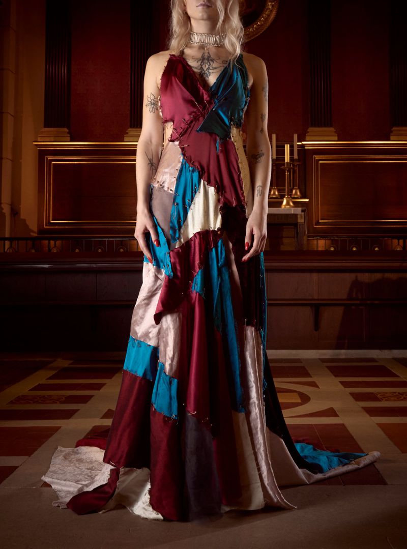 Woman in a patchwork-style evening gown, standing indoors with tile flooring and architectural details in the background, medium shot.