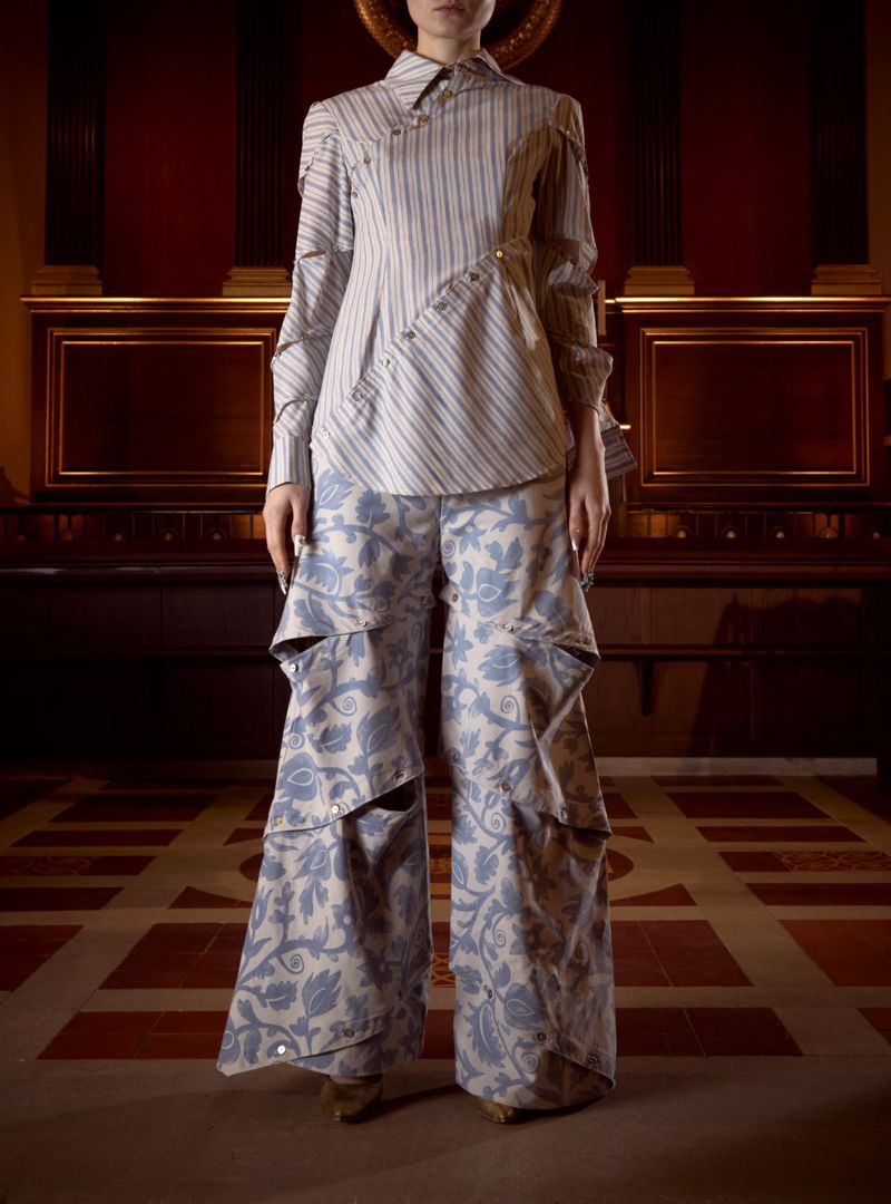 Model standing in an asymmetrical striped button-up shirt and wide-leg blue damask cutout trousers in a wood-paneled hall.