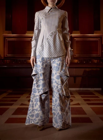 Model standing in an asymmetrical striped button-up shirt and wide-leg blue damask cutout trousers in a wood-paneled hall.