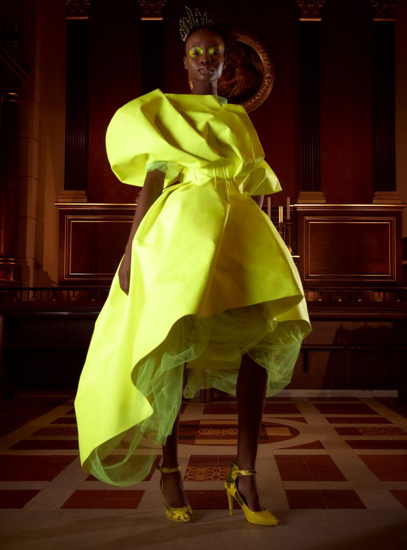 Woman in a sculptural neon yellow dress and matching heels stands in a dark, ornate interior with a tiled floor and decorative walls.