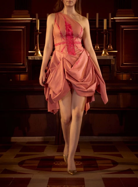 Young woman in an asymmetrical peach-colored dress and gold heels stands in an indoor space with dark wooden features and tile flooring.