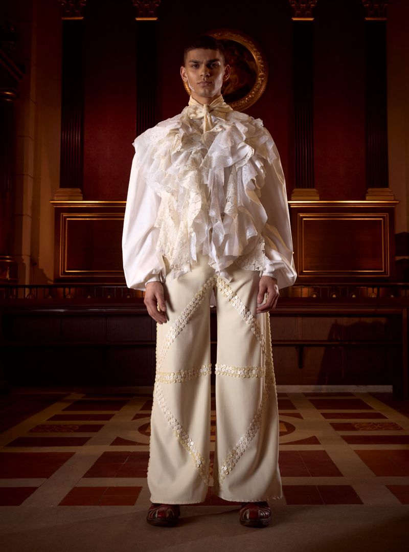 A stylish male model stands indoors with a dramatic ruffled shirt and embroidered pants, posing against a dark paneled background.