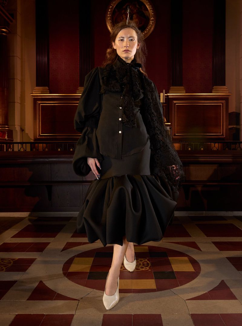 Elegant woman in black period-inspired fashion ensemble poses on tiled floor indoors with ornate architectural backdrop. High fashion editorial.