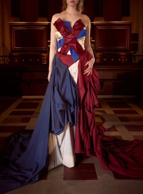 Woman in draped gown of red, white, and blue fabric, posing indoors with neutral expression in a dimly-lit location with tiled floors.