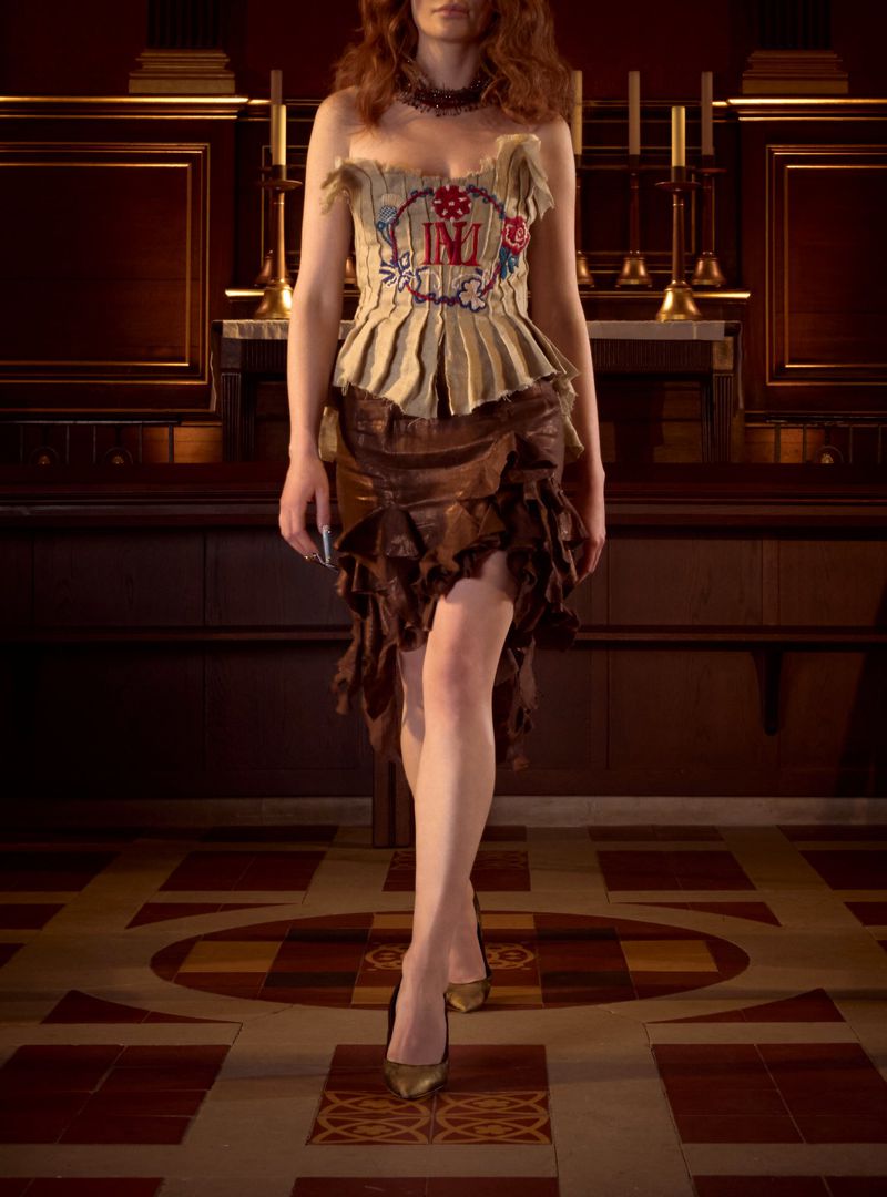 Woman in an embroidered top and brown skirt poses in a dimly lit room with a geometric floor, showcasing fashion and design.