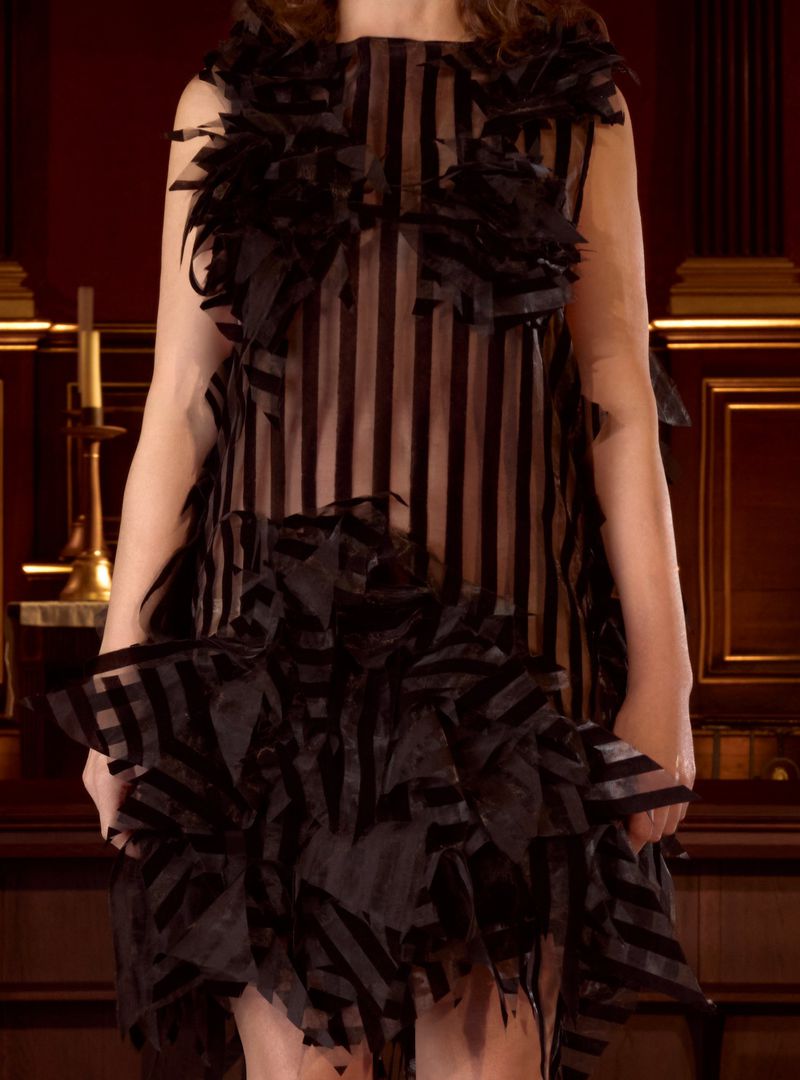 A woman models a black striped sleeveless dress with frills. The photo captures a full shot of the dress, set against a dark background.