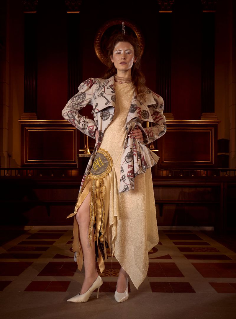 Adult female fashion model poses in a cream dress and coat with bicycle graphics indoors with dark wood panels and floors.