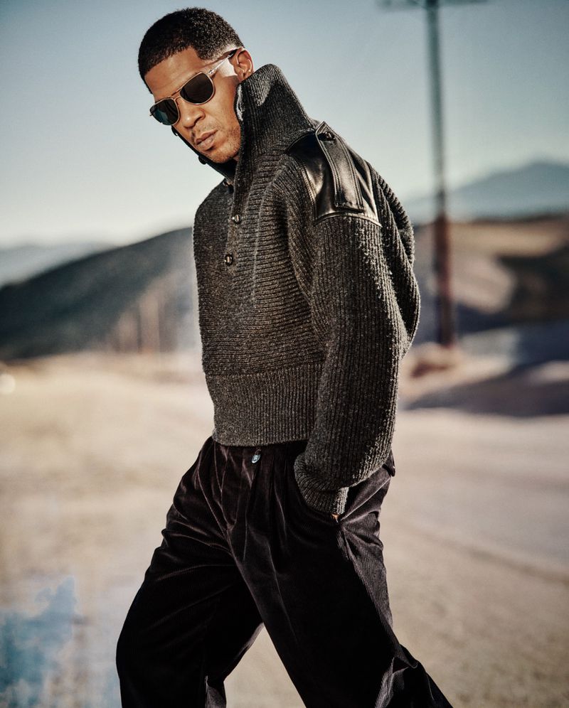 Kid Cudi walks outdoors wearing a high-collar grey knit sweater with leather shoulder panels, black trousers, and sunglasses.