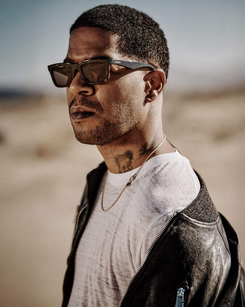 An adult male wearing sunglasses and a jacket stands against a sandy, outdoor background with clear skies in a medium portrait shot.