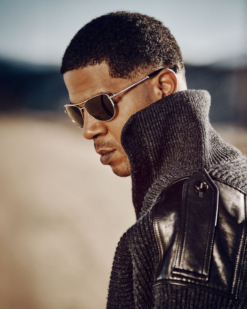 Portrait of a stylish adult male wearing aviator sunglasses and a grey sweater with a black leather shoulder piece, outdoors.
