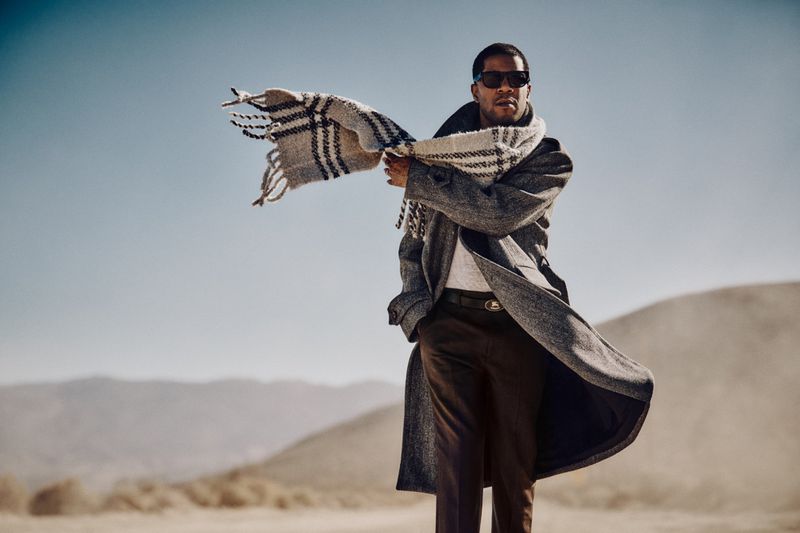 Adult male poses outdoors in a grey coat and checkered scarf against a desert landscape and clear blue sky on a sunny day.