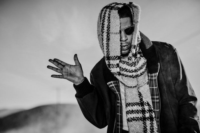 Kid Cudi in a plaid hooded scarf and jacket gesturing against an outdoor sky background