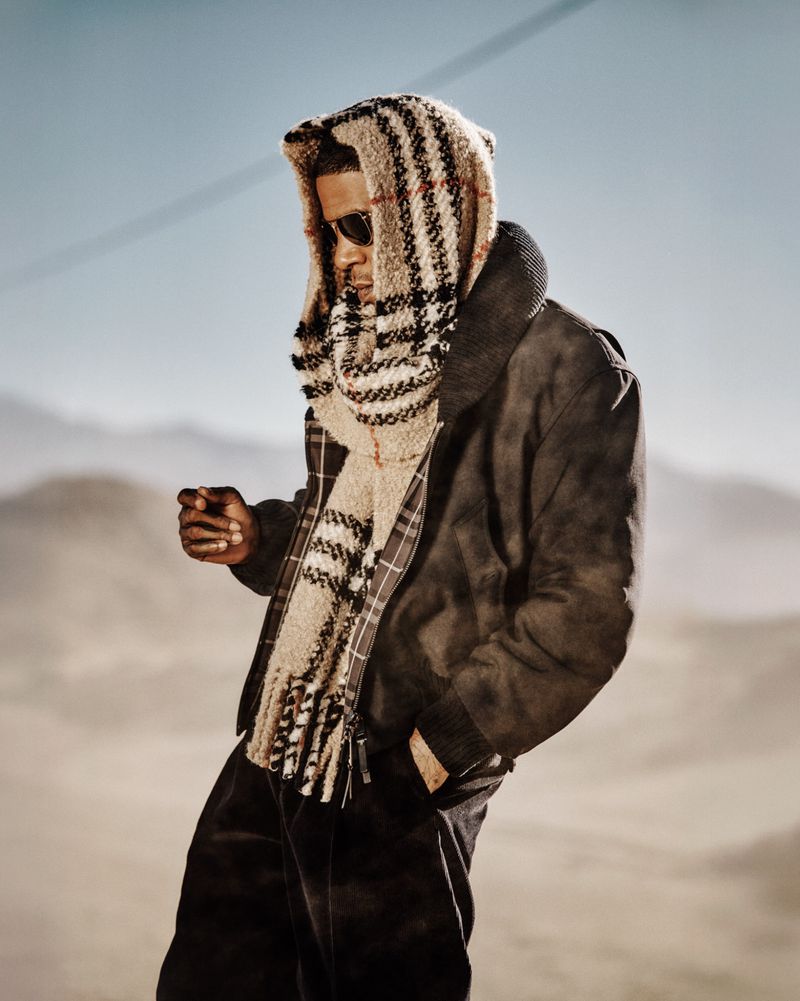 Kid Cudi wearing a hooded plaid scarf, sunglasses, and a dark brown jacket against a desert landscape.