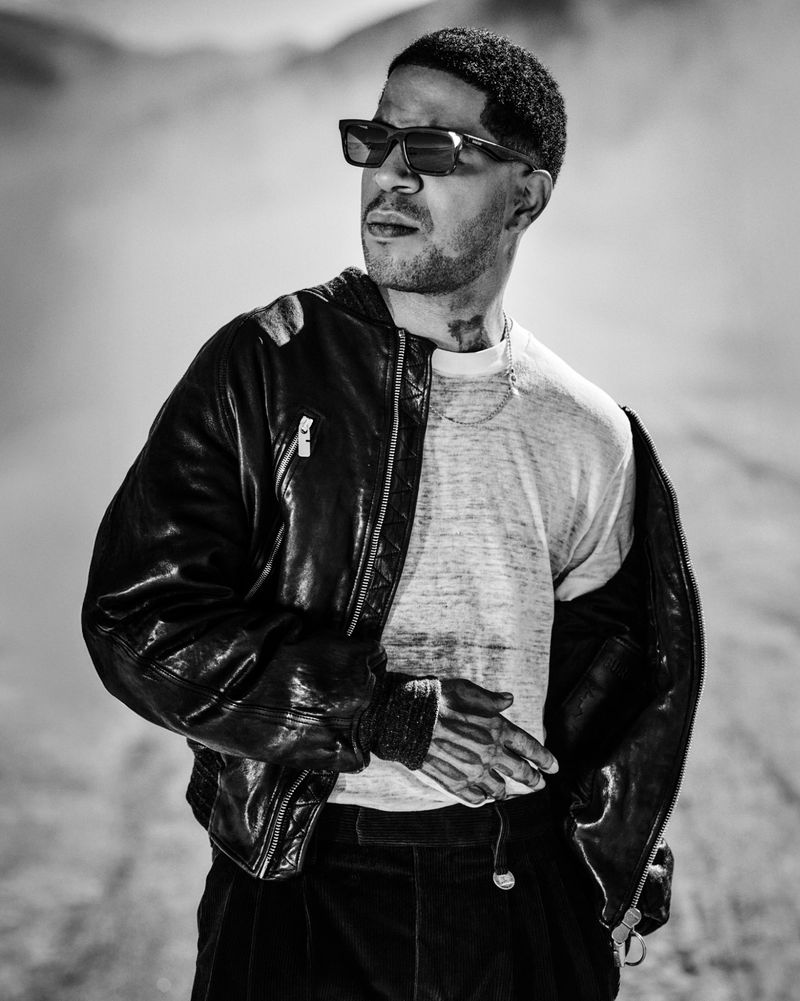 Black and white portrait of a man with sunglasses and a leather jacket in a dusty environment, looking off to the side.