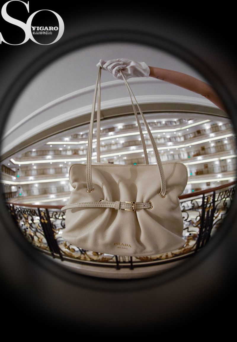 Cream-colored leather Prada handbag is displayed in front of an indoor balcony with railings, seen through a fisheye lens.