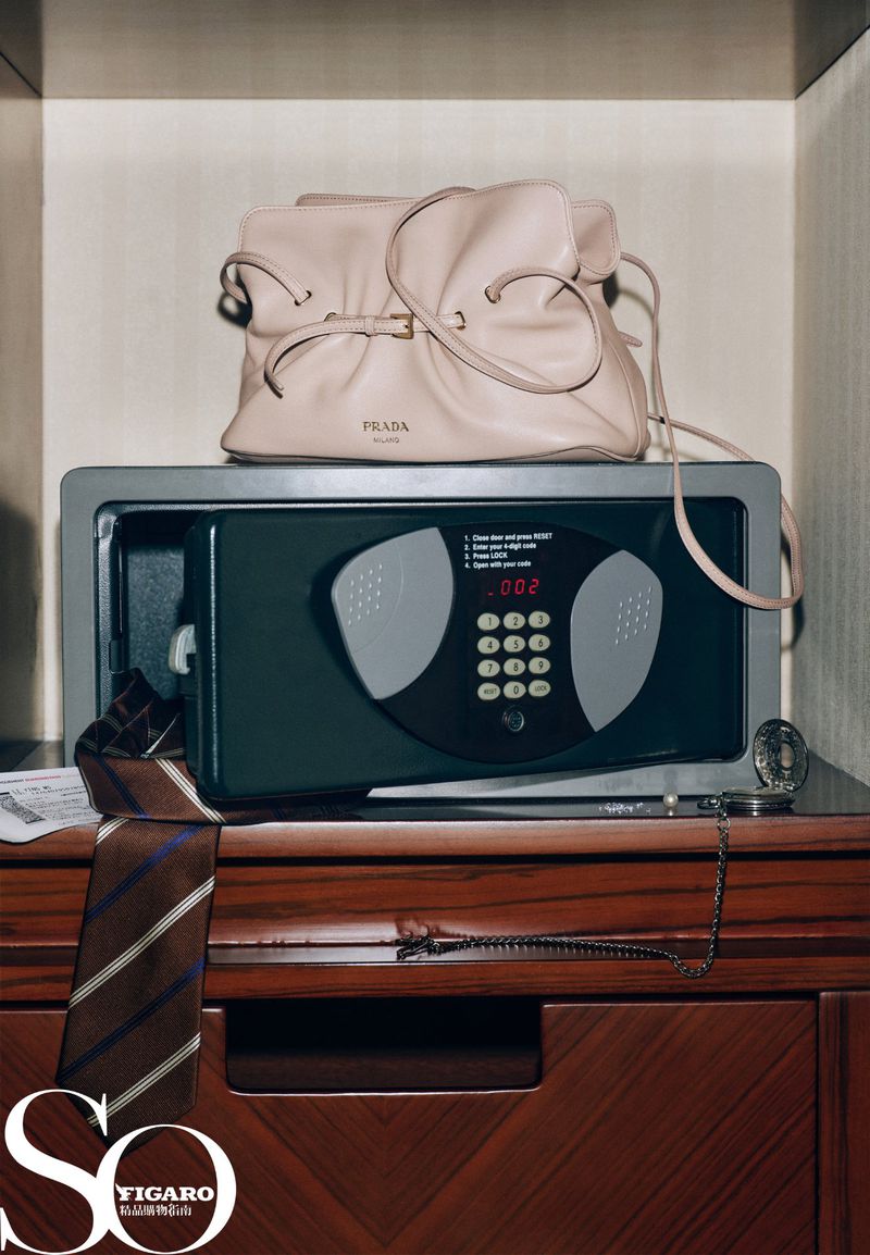Prada handbag sits on a gray safe atop a wooden bureau, accompanied by a tie, chain, and magazine, showcased in a studio shot.