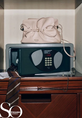 Prada handbag sits on a gray safe atop a wooden bureau, accompanied by a tie, chain, and magazine, showcased in a studio shot.
