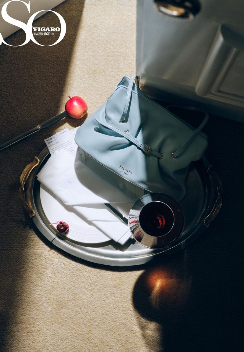Light blue leather handbag arranged on a silver tray with a white napkin, glass, and red cherry under angled sunlight.