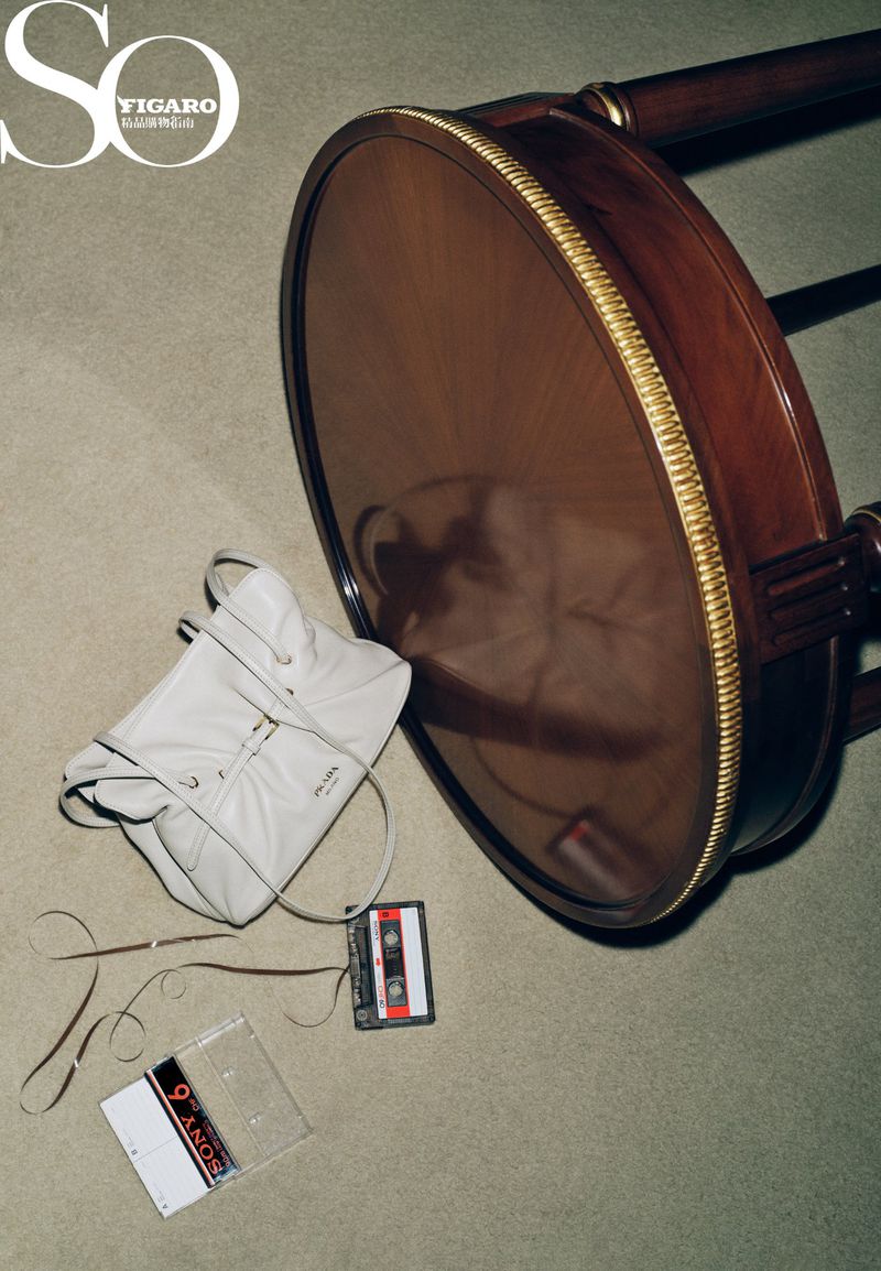 Still life photograph showcasing a tan leather shoulder bag, cassette tape, and tape case on a neutral carpet, beside a round table.