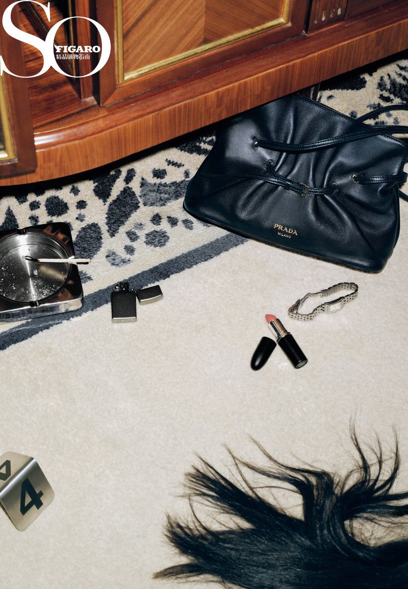 High-angle shot featuring a black Prada purse, lipstick, lighter, ashtray, watch, hair strands, and dice on a patterned carpet.