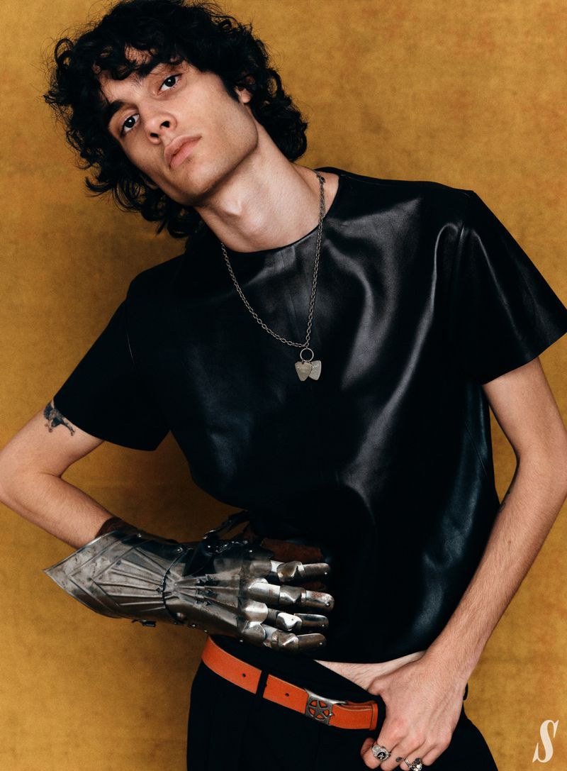 Adult male poses indoors with a metal gauntlet, wearing a black shirt, orange belt, and silver necklace against a gold backdrop.