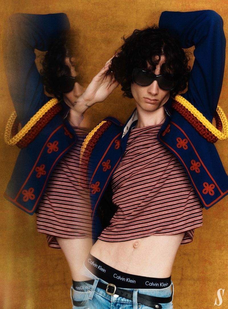 Fashion editorial portrait of adult male model with sunglasses wearing a horizontally striped t-shirt, jacket, and jeans in a studio.