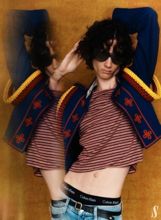 Fashion editorial portrait of adult male model with sunglasses wearing a horizontally striped t-shirt, jacket, and jeans in a studio.