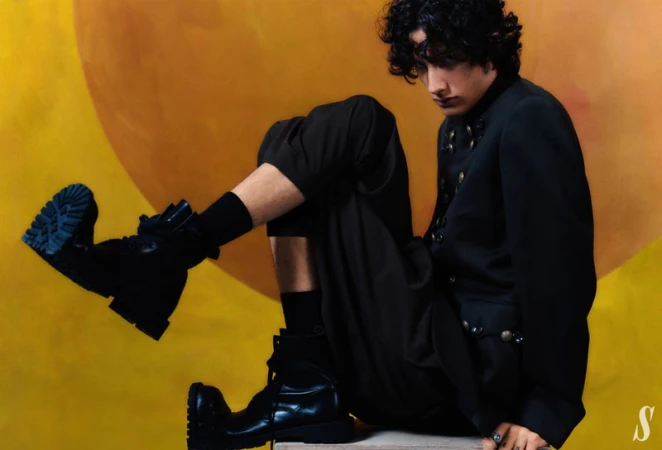 Fashionable young adult in black military-style jacket sits with black combat boots and yellow painted backdrop.