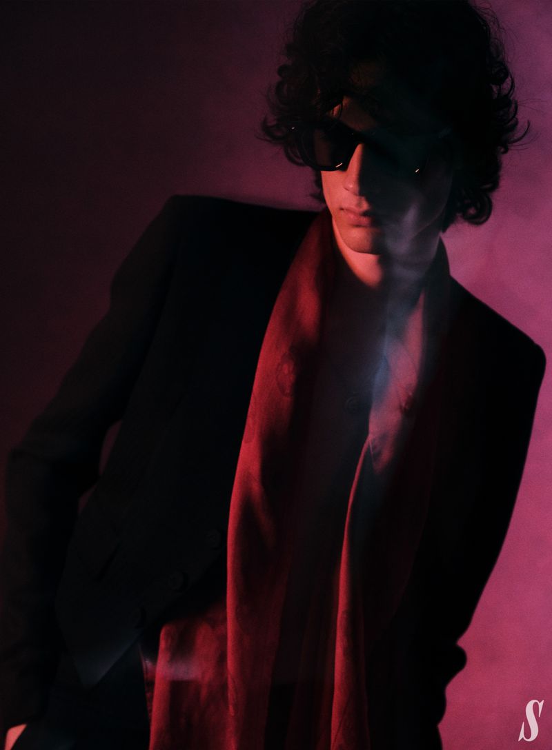 Portrait of an adult male with curly hair wearing sunglasses, a black suit jacket, and a red scarf, set against a dark pink backdrop.