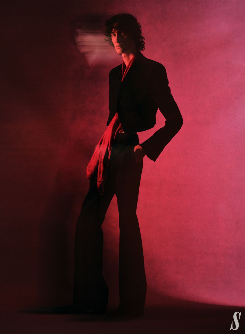 A slender adult male fashion model with curly hair poses in a low-lit photo studio wearing a cropped black blazer and wide-leg pants.