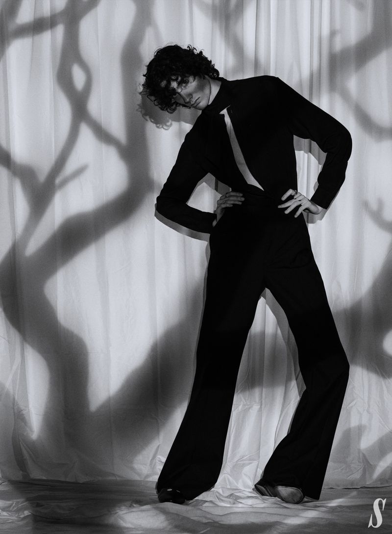 Monochrome studio portrait of a slender adult wearing a black jumpsuit, standing in front of a textured white backdrop with tree branch shadows.