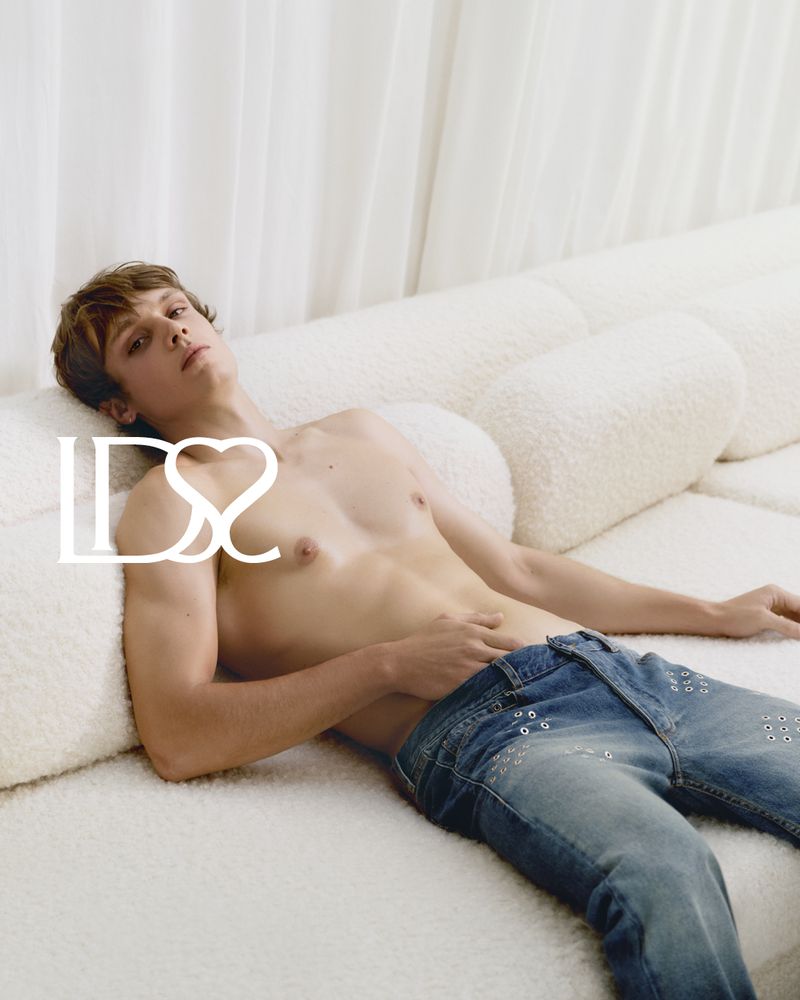 Young adult male poses on a plush white sofa in a light-filled interior wearing blue jeans, capturing a blend of casual and stylish aesthetics.
