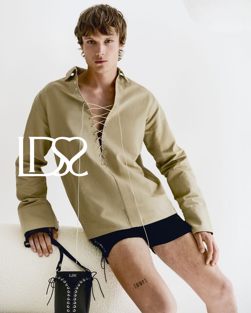Fashionable male model poses indoors in an oversized tan shirt with lace-up detail, black shorts, and a stylish bucket bag with eyelets.