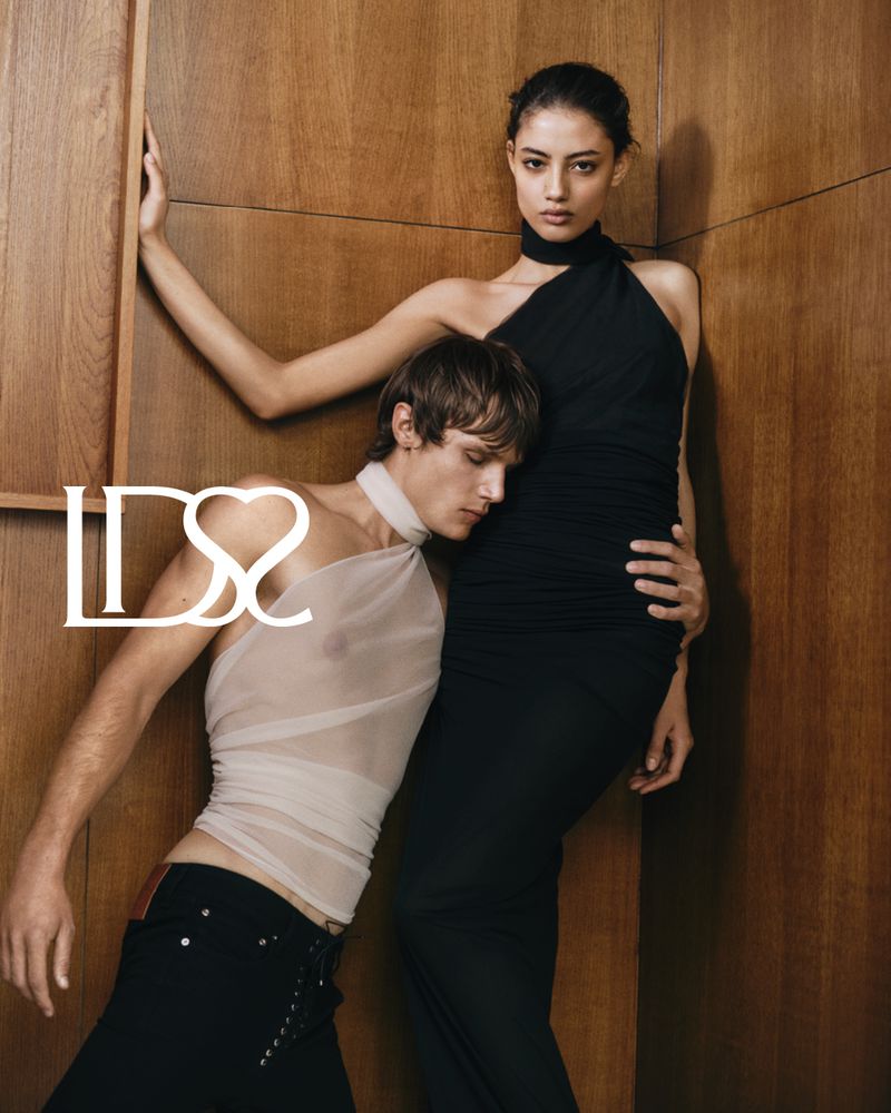 Fashion shot of a woman in black dress and a man in sheer top, posing against a wood wall. Moody fashion, dark colors.