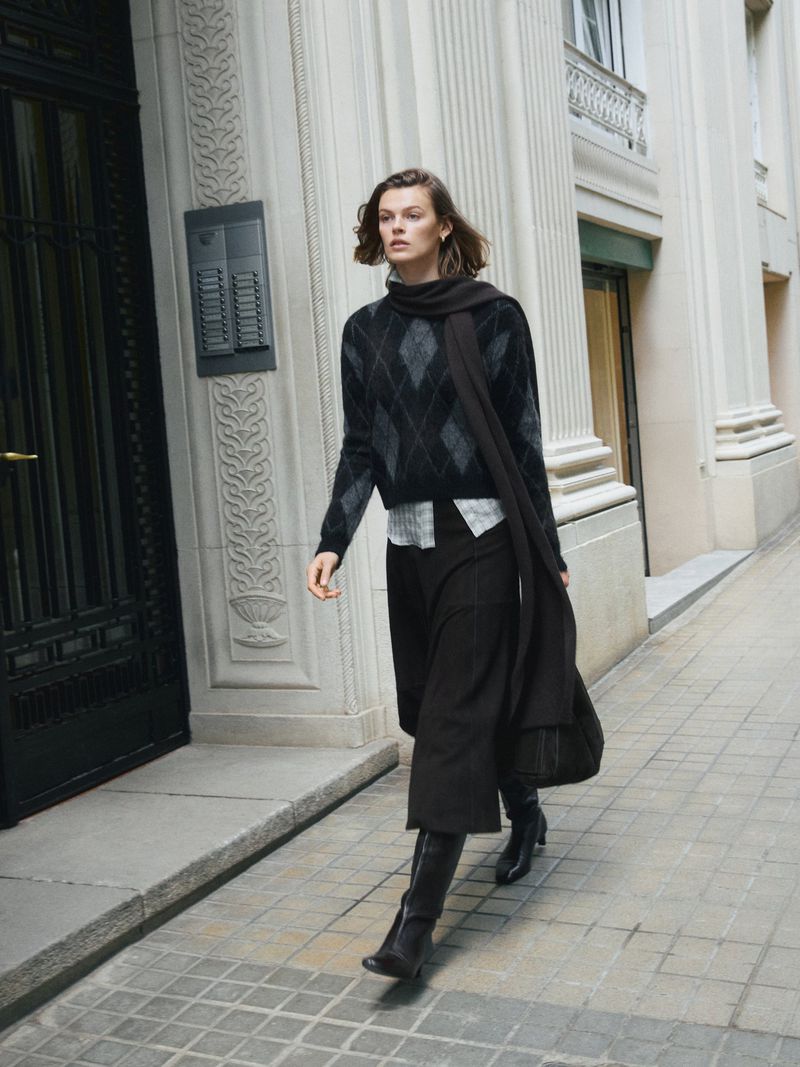 Fashionable woman in an argyle sweater and midi skirt walks along an urban sidewalk near a building with architectural details.