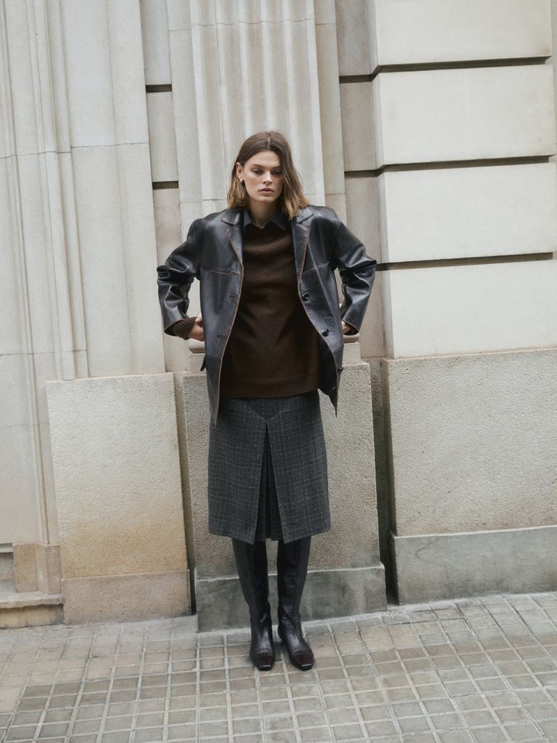 A woman in a black jacket, brown sweater, gray skirt, and black boots stands against a light gray wall outdoors, gazing downward.