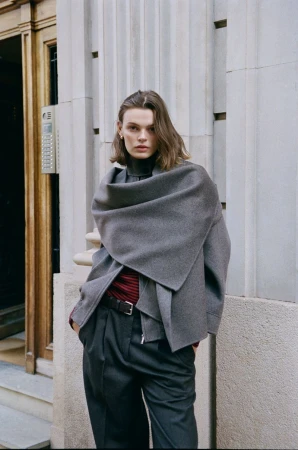 Fashionable adult female poses outdoors wearing a grey scarf-jacket, turtleneck top, belted pants, and an earring, against a textured wall.