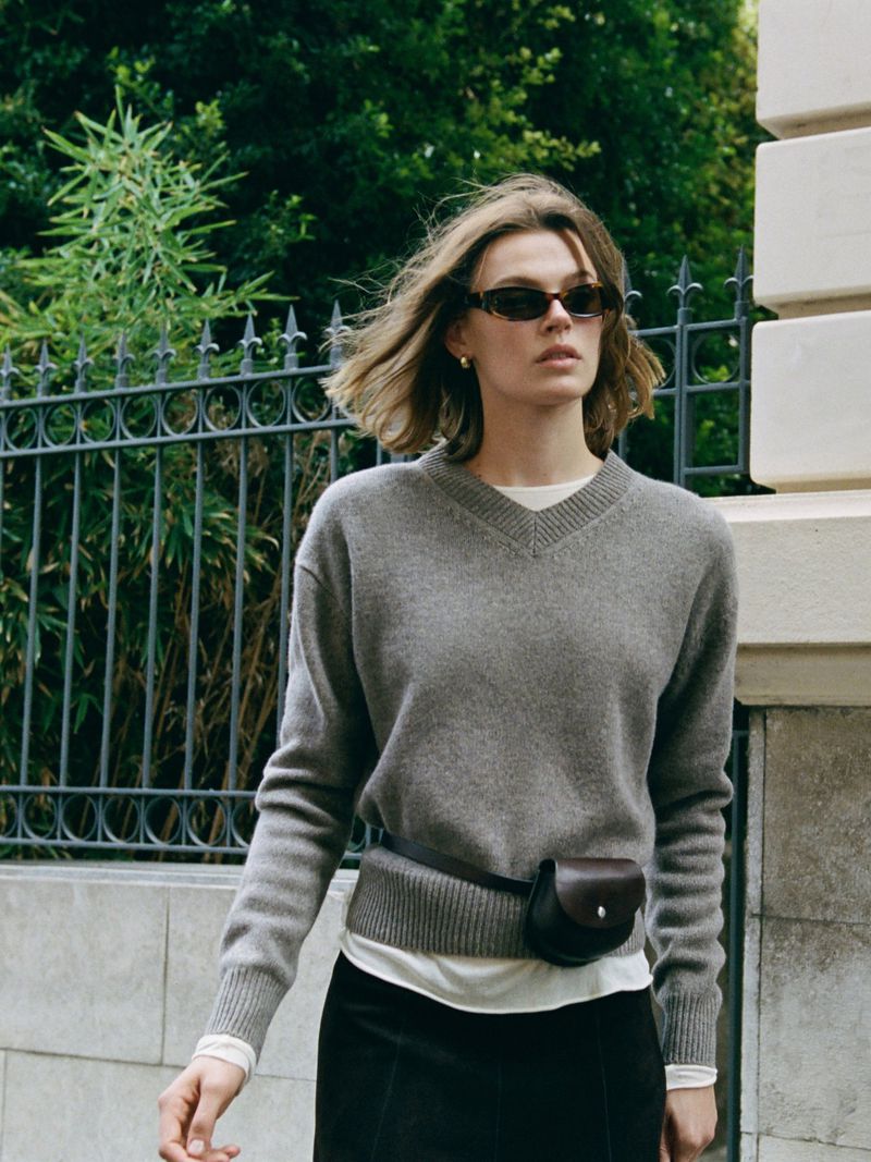 Model wearing a grey V-neck sweater and sunglasses outdoors against a black metal fence and stone building wall