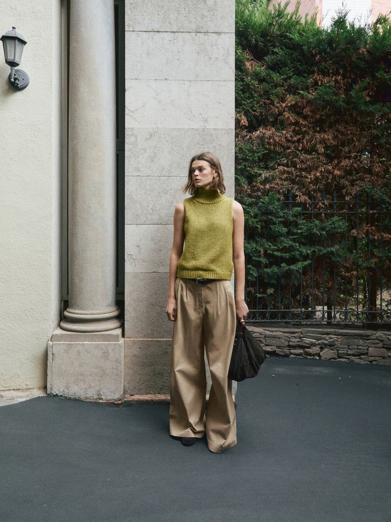 A full outdoor shot of a woman in a knit top and wide-legged pants posing next to a light-colored stone column and leafy green trees.