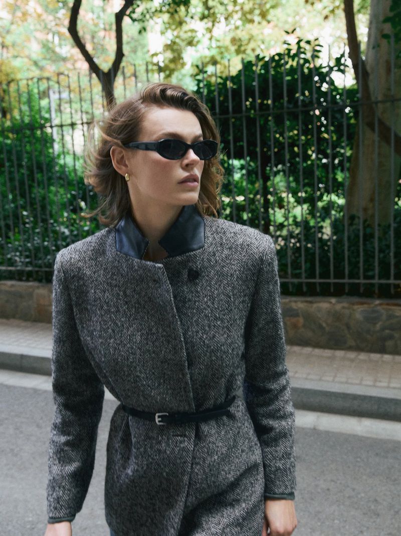 Woman in gray coat and sunglasses walks on city street with trees in background on a sunny day; fashion, style, and outdoor scene.