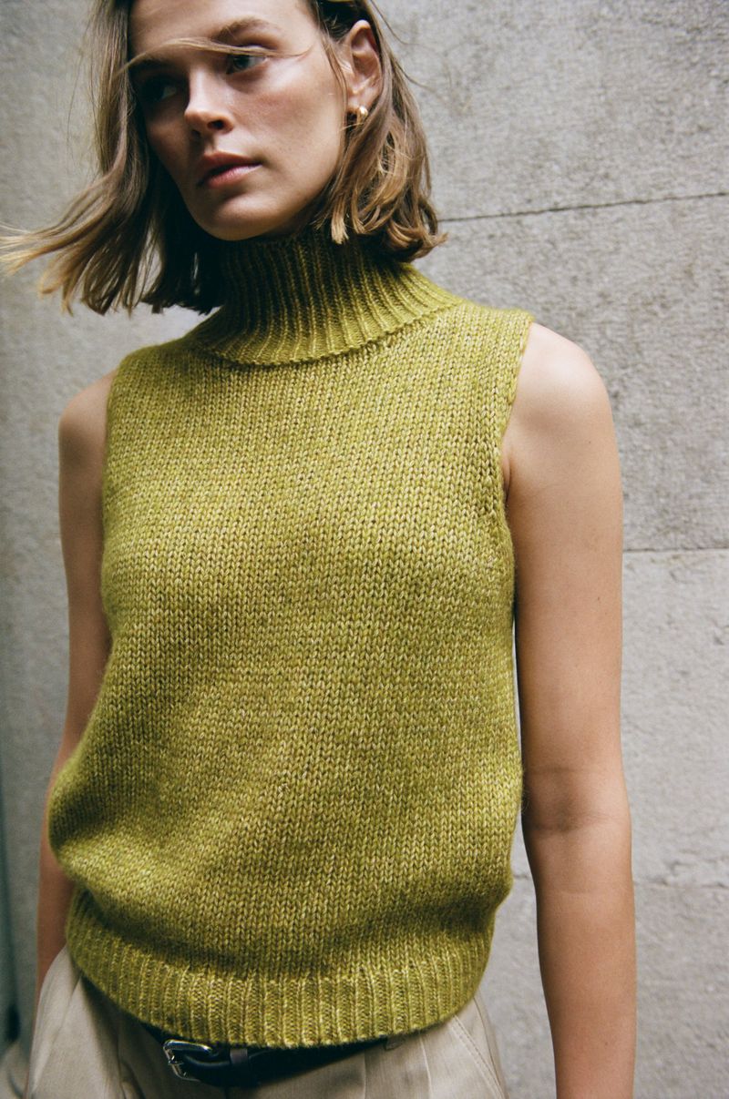 Model Cara Taylor wearing a sleeveless chartreuse knit turtleneck vest against a textured stone wall.