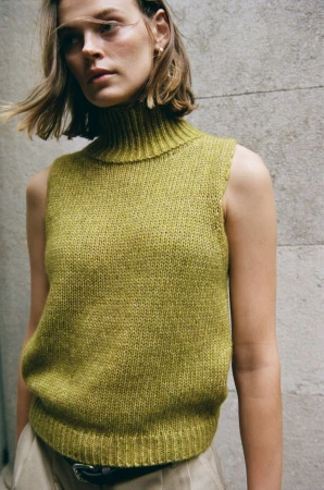 Model Cara Taylor wearing a sleeveless chartreuse knit turtleneck vest against a textured stone wall.