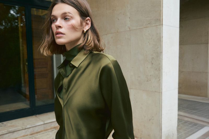 Portrait of a woman in a green satin shirt, standing against a textured beige wall, looking slightly off camera in natural light.
