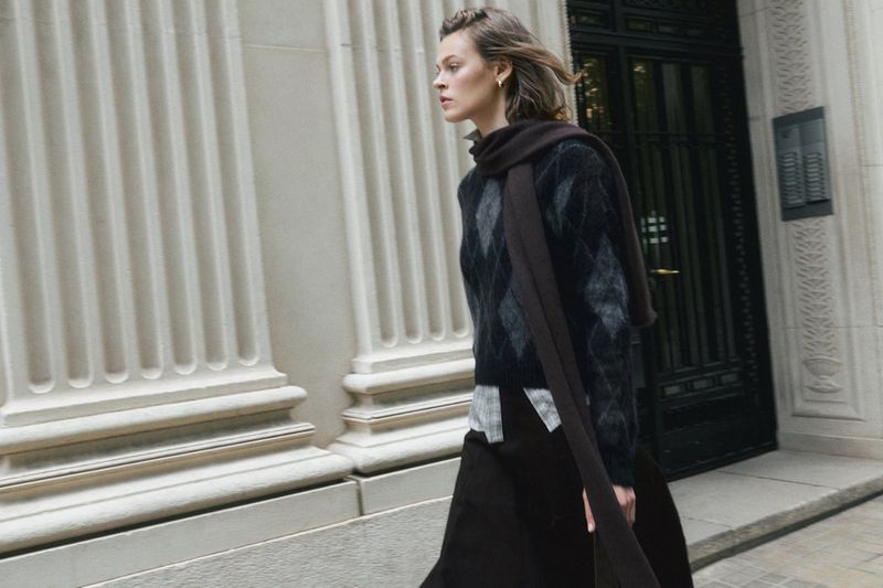 Woman in argyle sweater walks past stone building on city street. Brown scarf drapes over her shoulder. Elegant, neutral tones.