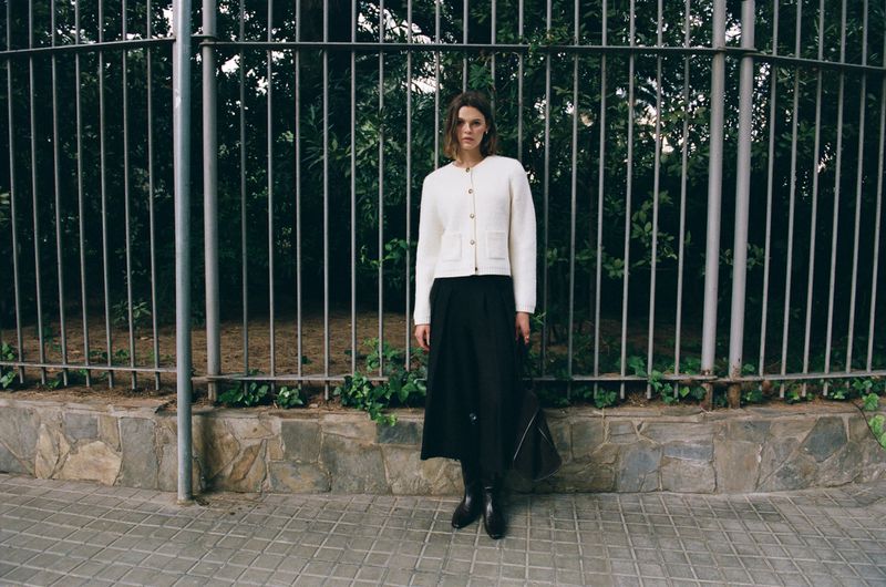 A woman in a cream cardigan and black skirt poses in front of a metal fence in an outdoor urban setting during the daytime.