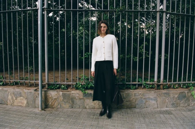 A woman in a cream cardigan and black skirt poses in front of a metal fence in an outdoor urban setting during the daytime.