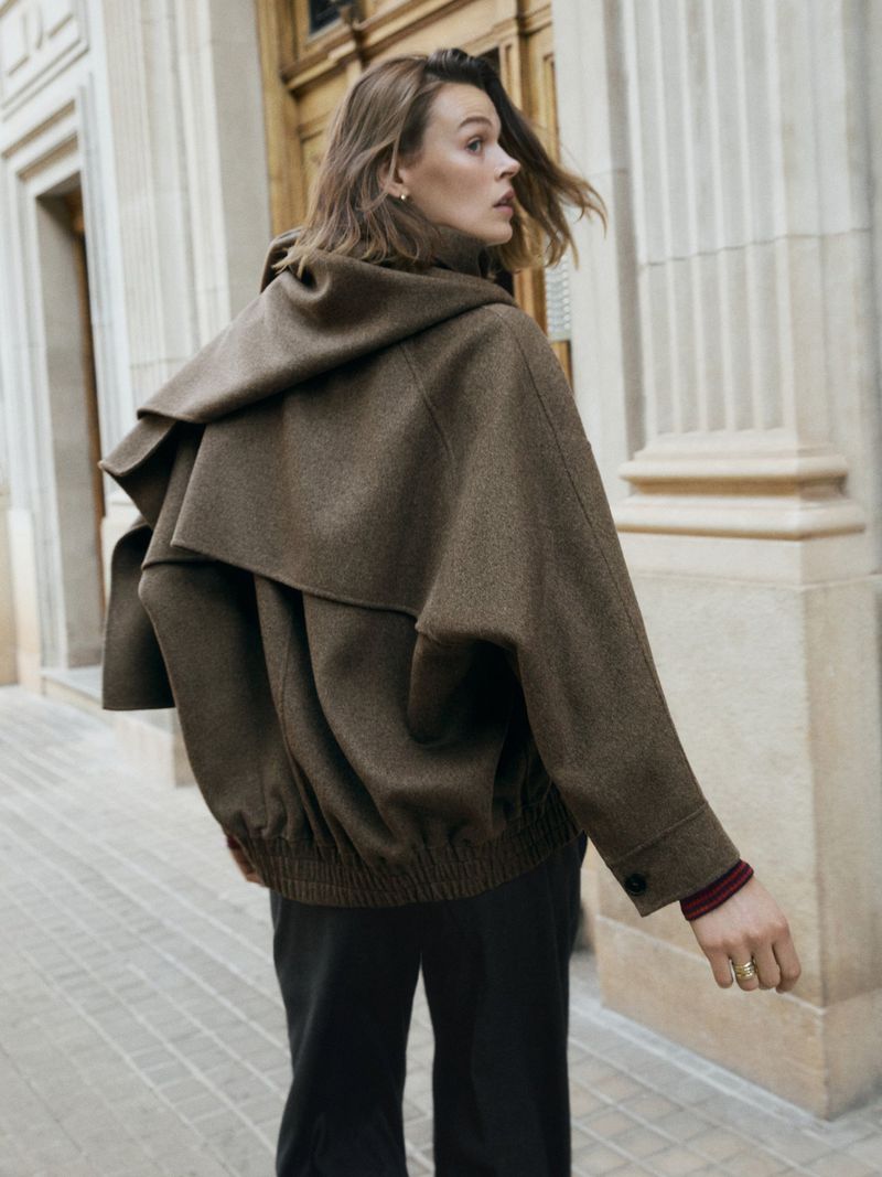 Woman wearing a large brown hooded jacket walks past an exterior wall with arched door frame and gray stone sidewalk in daytime.