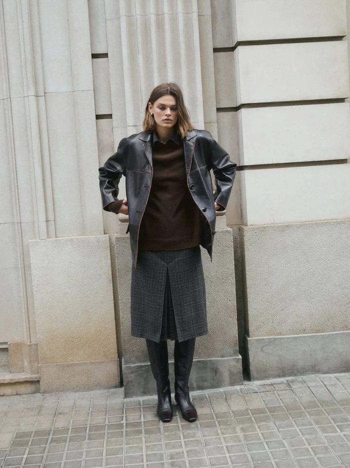 Model Cara Taylor in a dark leather jacket and grey pleated skirt standing outdoors against a stone building for Massimo Dutti