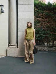Model Cara Taylor wearing a green sleeveless knit turtleneck and pleated beige wide-leg trousers standing outdoors by a stone pillar.