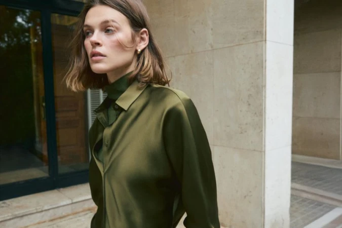 Model Cara Taylor wearing an olive green satin button-up shirt outdoors against a limestone architectural wall.